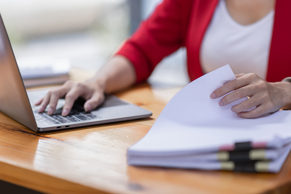 A woman works on laptop computer while flipping through a stack of documents