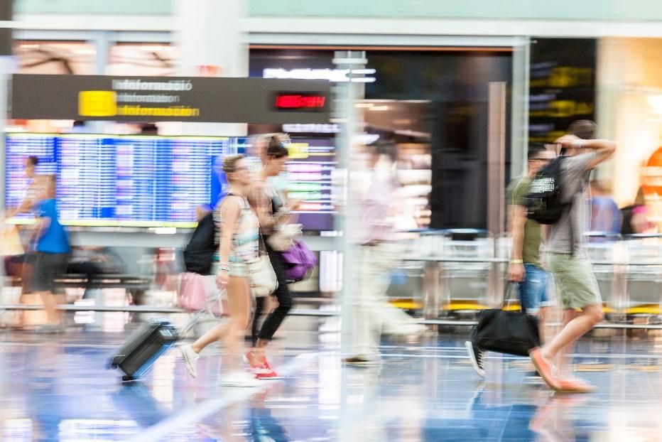 Colorful, blurred image of airport travellers walking by a departures board depicting the busy, frantic pace of airports that possible innovations and solutions need to consider.