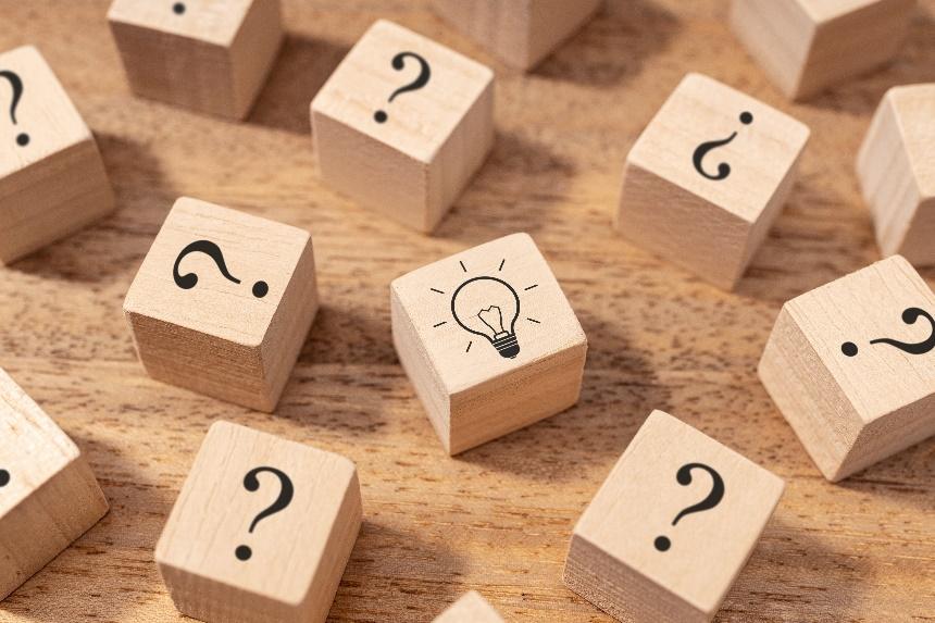 One square wooden block with a lightbulb drawing sits among a field of cubes marked with question marks to depict an innovative idea popping up amid an abundance of questions.